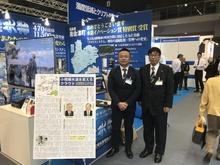 青いパネルの前に立っている2名の男性の写真