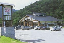 駐車場入口に看板があり、車が数台駐車されている奥に茶色い屋根で白い壁の山口温泉道の駅きらら289の建物が写っている写真