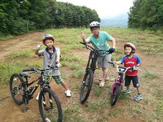 樹々に囲まれた広場で男性1人と男の子2人がマウンテンバイクの横に立ち笑顔で写っている写真