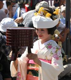 丸髷の髪をして白の着物にピンクの帯をした女性が七行器を持っている写真