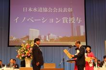 「日本水道協会会長表彰 イノベーション賞授与」と書いてあるプロジェクタースクリーンの前で1人の男性が賞状を読み上げ、向かい側にスーツを着た男性が立っている写真