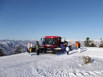 雪で真っ白な山頂に停まっている赤い雪上車と、その周りに6名の方々が山頂を歩いている写真