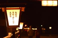 参道に「祇園祭意地で押し出す大屋台」と書かれた行燈が設置され、暗闇に灯りが灯っている写真