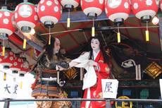 赤い提灯の飾られた神輿の中で、鎧を身にまとった男性と、赤い着物を着た女性が演技をしている写真